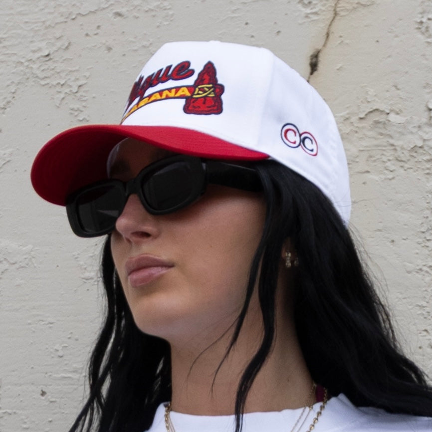 Person wearing a white and red cap with branding, sunglasses, and a white shirt against a neutral background.