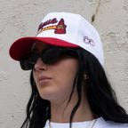 Person wearing a white and red cap with branding, sunglasses, and a white shirt against a neutral background.